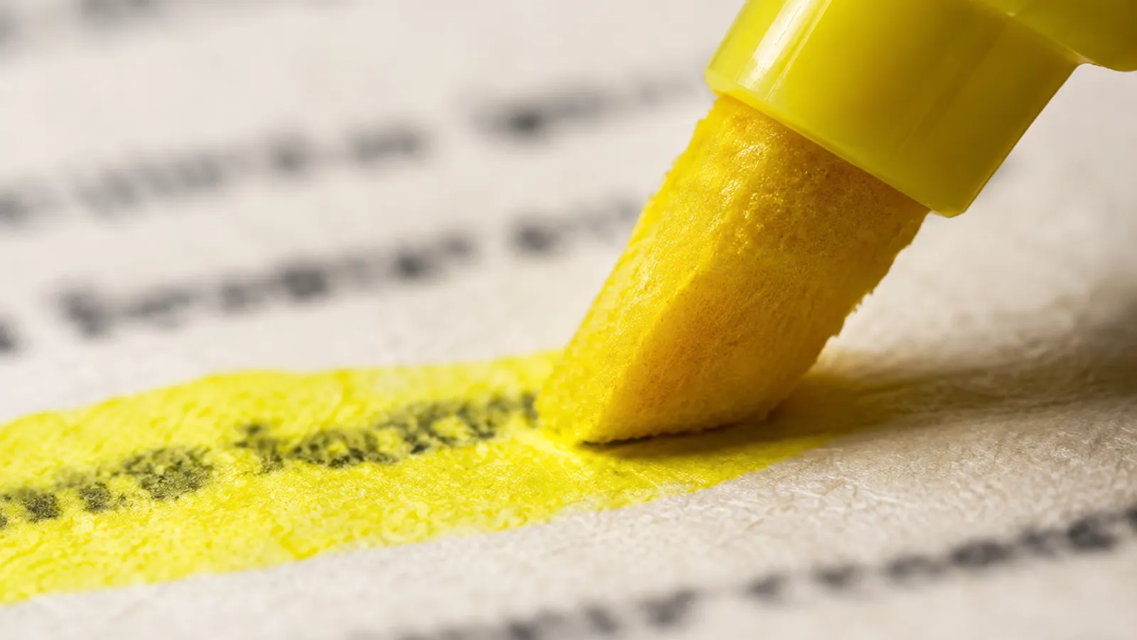Extreme close-up of a yellow highlighter tip mid-stroke across a printed line, the ink soaking into the paper fibres and the text out of focus.